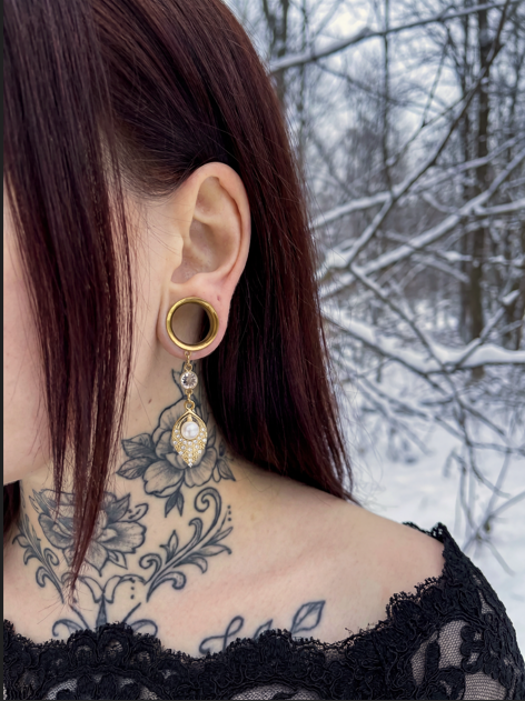 Boucles d'oreilles dorées avec perle nacrée et strass, portées par une femme avec cheveux roux foncés et tatouages, dans un décor enneigé
