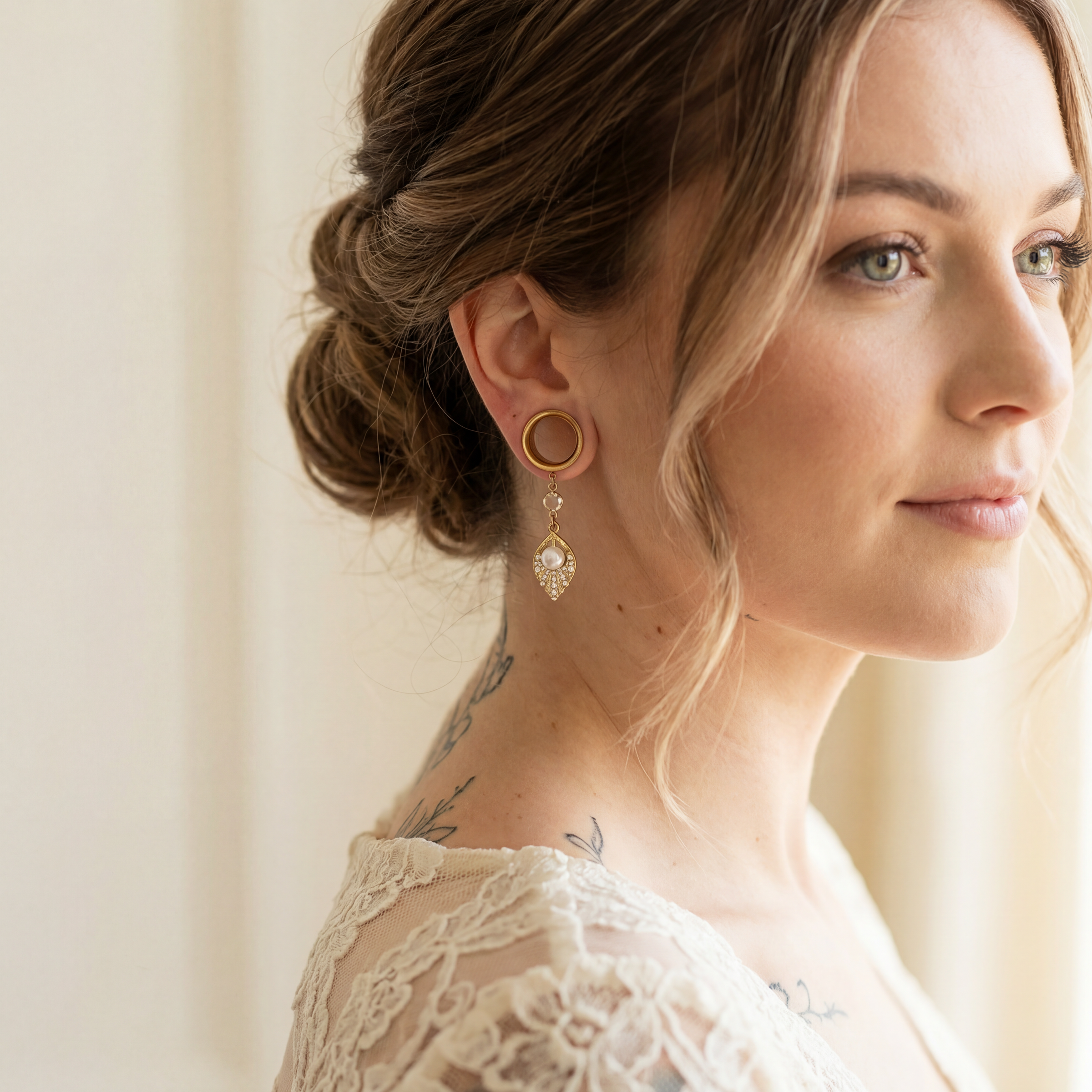 Boucles d'oreilles dorées avec perle nacrée et strass, portées par une femme avec cheveux châtains clairs et tatouages, en intérieur lumineux