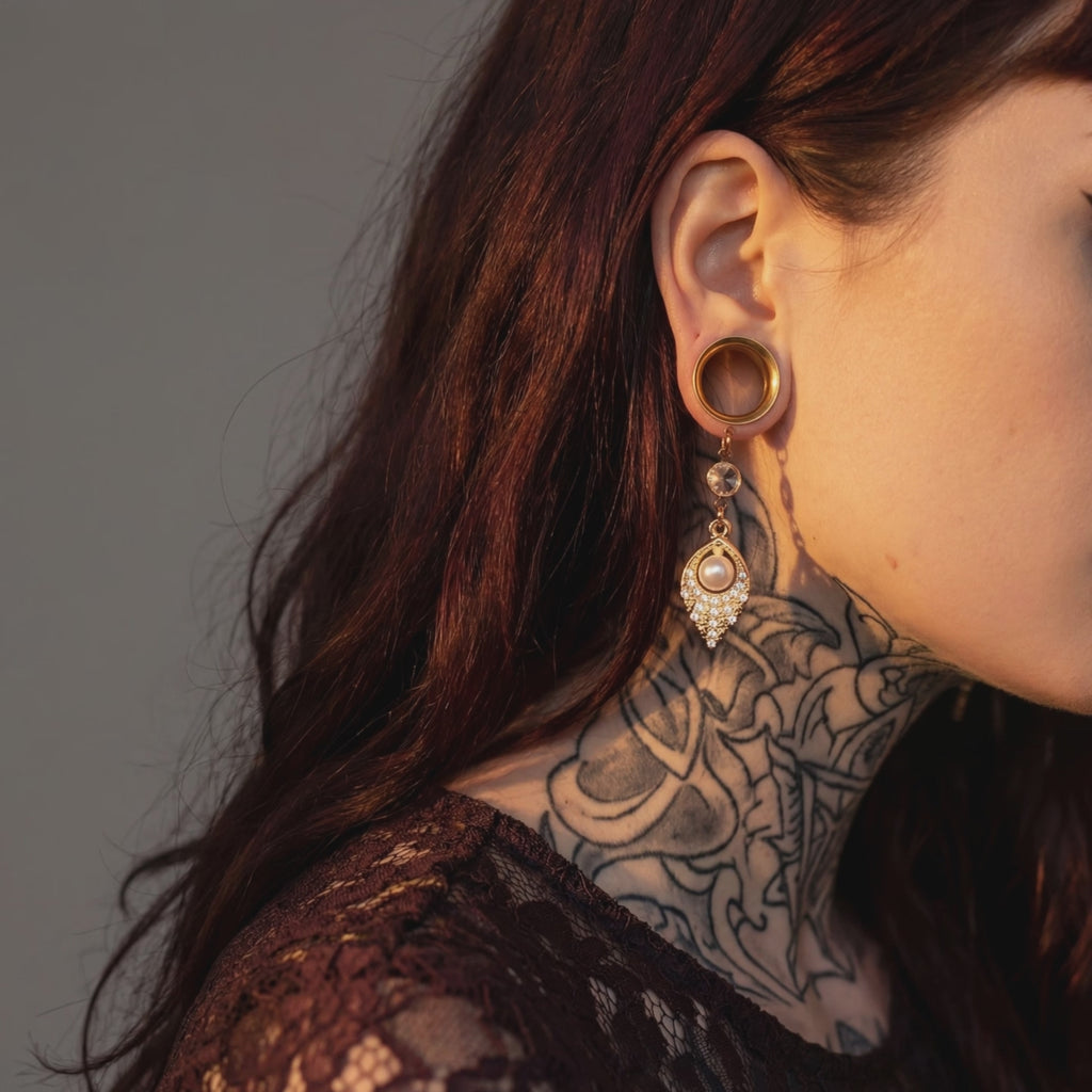 Boucles d'oreilles pendantes dorées avec perle nacrée et strass scintillant, portées par une femme avec cheveux roux et tatouages, en intérieur
