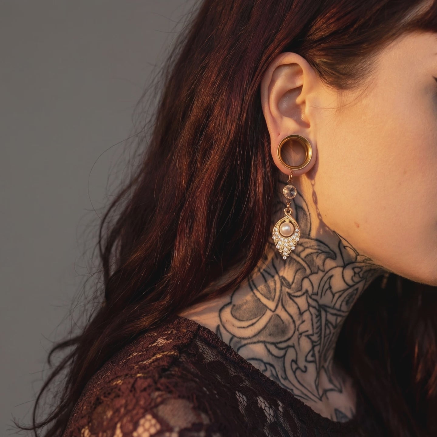 Boucles d'oreilles pendantes dorées avec perle nacrée et strass scintillant, portées par une femme avec cheveux roux et tatouages, en intérieur
